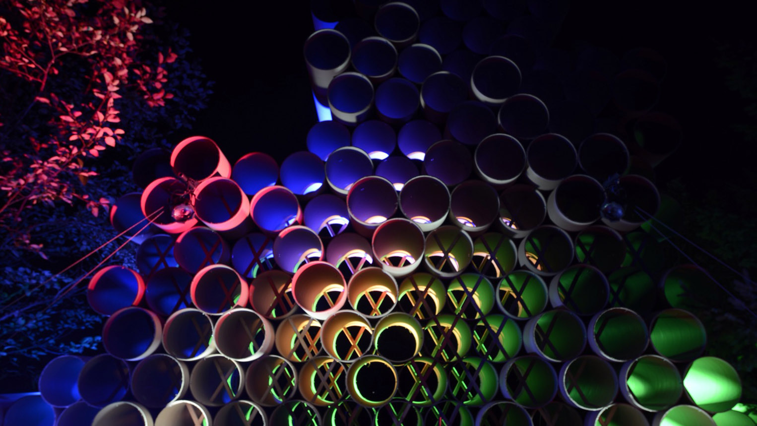 Night shot of rainbow lit mountainous 15 foot wall of horizontally stacked large cardboard tubes with ends towards camera